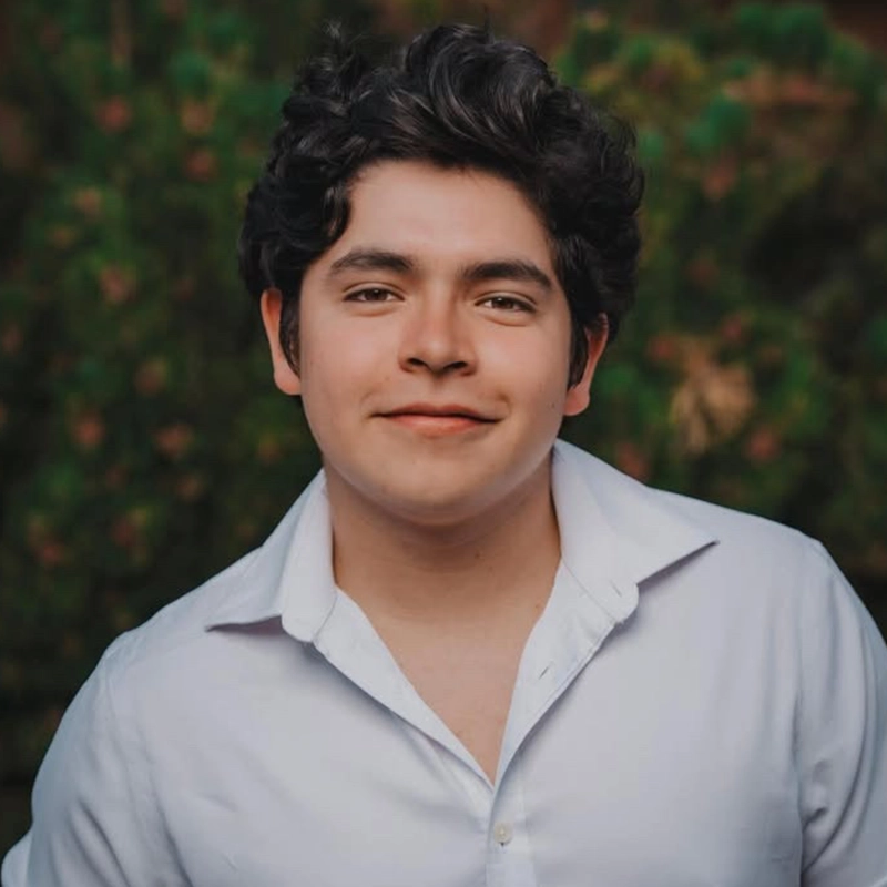 Sergio Guerra, a young man with wavy dark hair and fair skin, smiles gently. He wears a slightly unbuttoned white collared shirt. The blurred outdoor background features green foliage, while soft, natural lighting enhances the scene.
