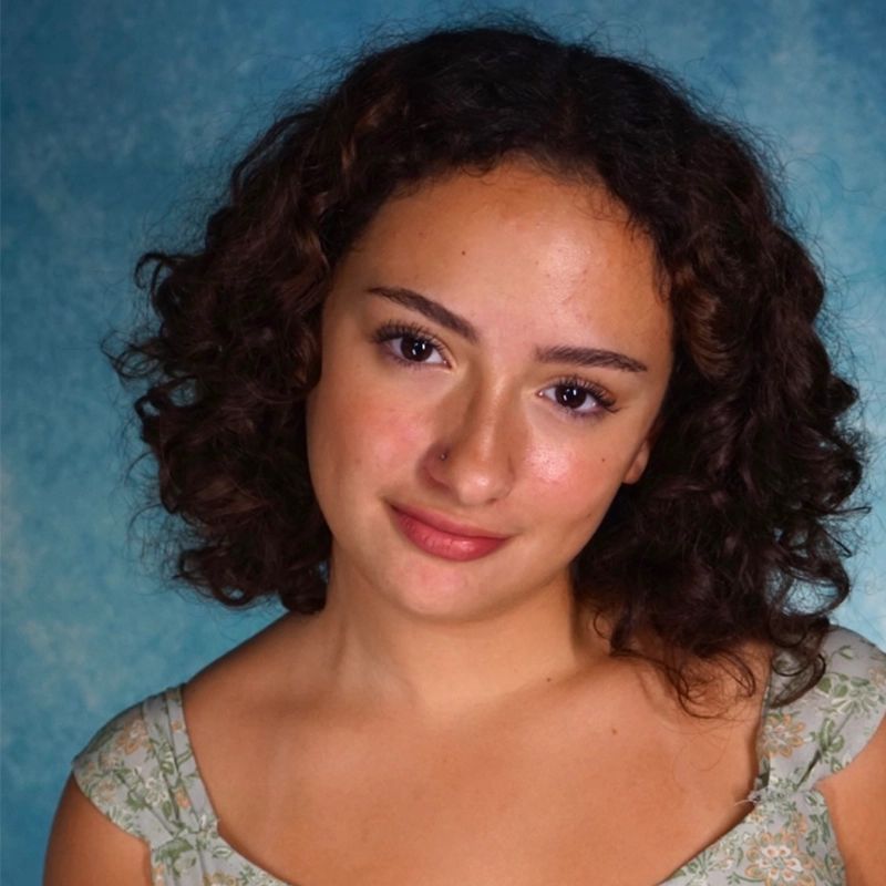 A young woman with curly brown hair and light skin smiles softly at the camera. She wears a light green, patterned top with cap sleeves. The studio portrait has a mottled blue background, reminiscent of Sergio Guerra’s distinctive style.