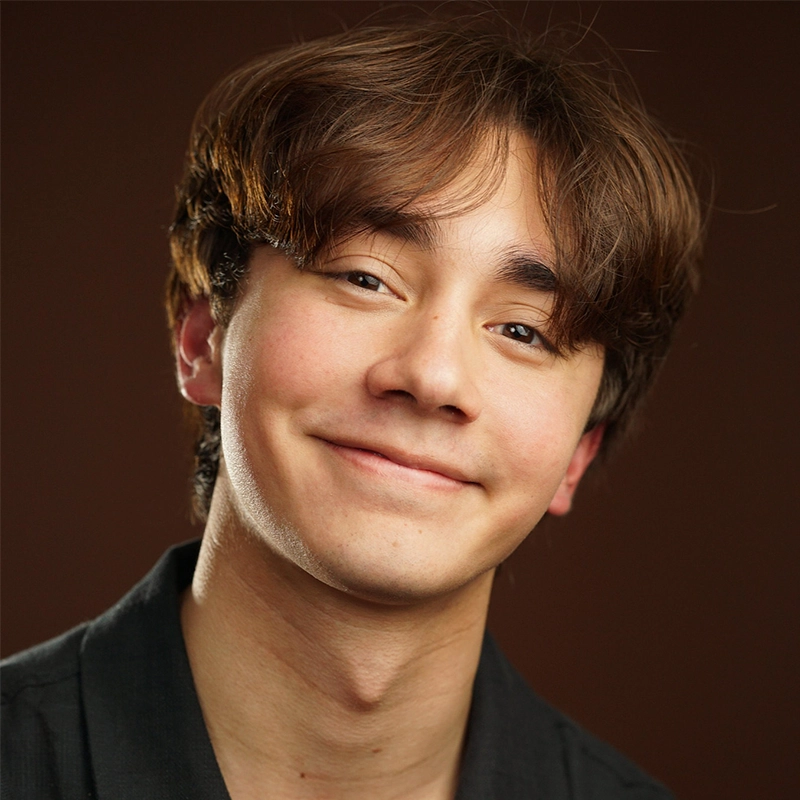 Jaeden Riley Juarez, a young person with light skin and medium-length brown hair, smiles gently. Wearing a dark shirt against a plain, dark background, they look directly at the camera as soft lighting highlights their features.