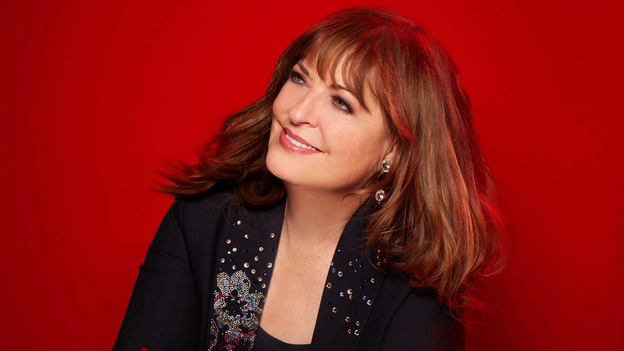 Ann Hampton Callaway - Show Art A woman with medium-length brown hair and bangs smiles while looking up to her left, echoing the joyful spirit of Ann Hampton Callaway’s “Here’s To Life.” She wears a black jacket with sparkling embellishments and dangling earrings, set against a vibrant solid red background.