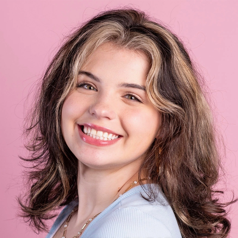 A young woman, Olivia Luzquinos, with wavy brown hair featuring light blonde highlights smiles at the camera. She wears a light blue top and a gold necklace. The solid light pink background adds to the cheerful, friendly mood.