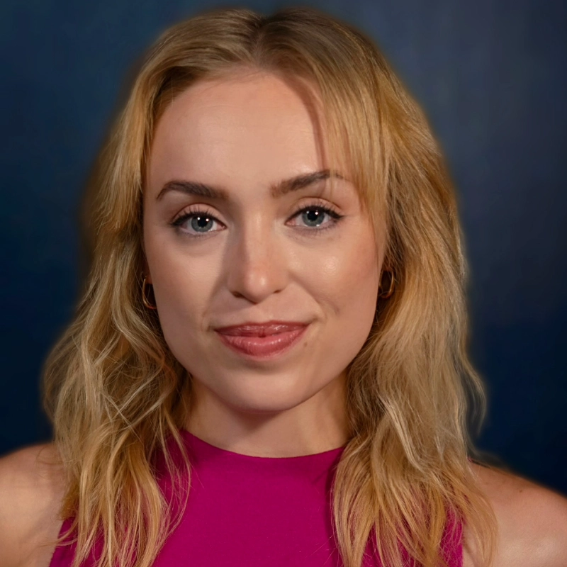 A woman with wavy blonde hair, light skin, and blue eyes smiles softly. She wears a sleeveless magenta top and small hoop earrings. The softly blurred blue background gives a serene feel to this Josiah Jennings-inspired portrait. Her makeup is natural, highlighting her features.