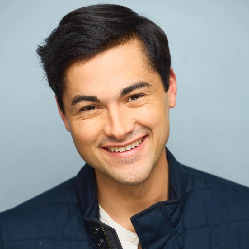 A young man with short dark hair smiles warmly at the camera. He is wearing a navy blue jacket over a white shirt and is posed in front of a plain light blue background. His expression is friendly and inviting.