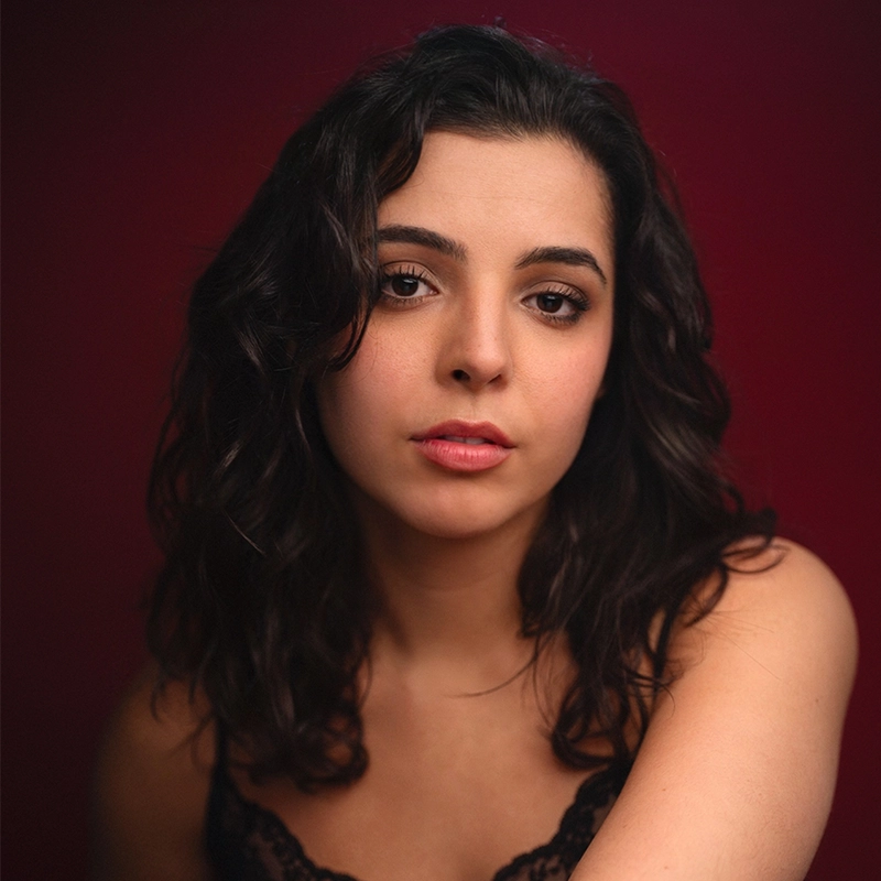 Bekah Zornosa, a young woman with wavy dark hair and light skin, gazes softly at the camera. She wears a black lace top and minimal makeup. The deep red background and gentle lighting create an intimate mood.