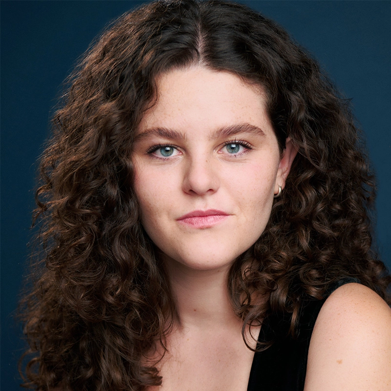 A young woman with fair skin and blue eyes looks directly at the camera. Olivia Luzquinos has long, thick, dark brown curly hair and wears a sleeveless black top. The solid dark blue background highlights her calm and confident expression.
