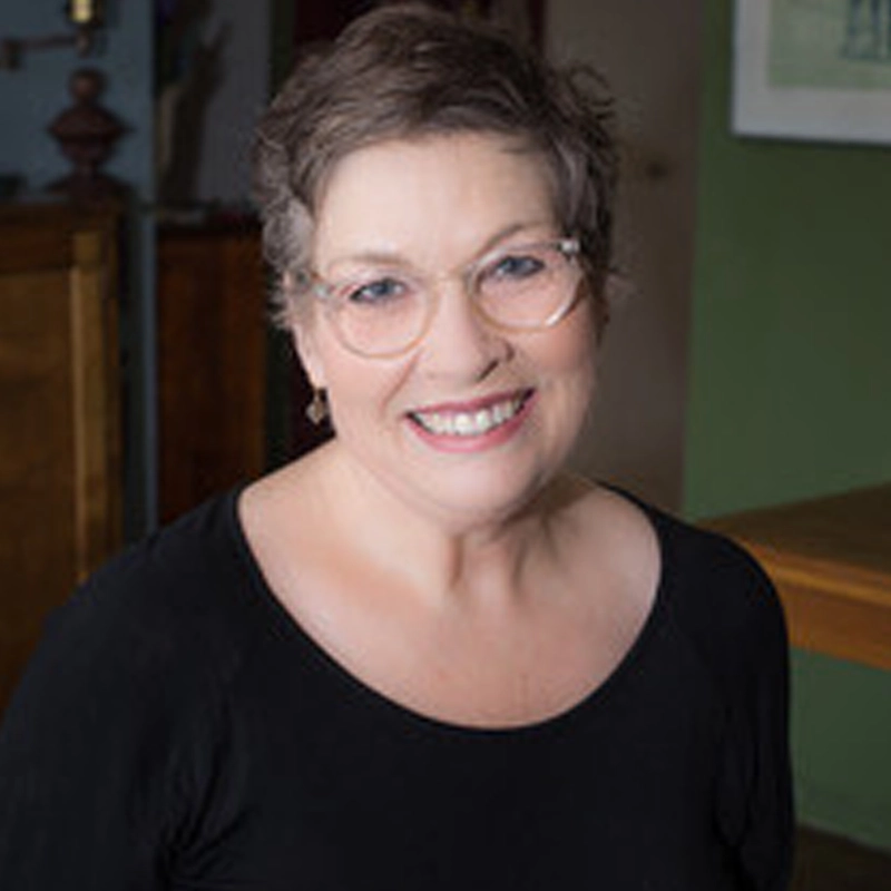 A smiling middle-aged woman, identified as Laura Bergquist, with short, curly brown hair and glasses wears a black top. She is indoors in a warmly lit room with green and wooden decor visible in the background, exuding a friendly and welcoming demeanor.