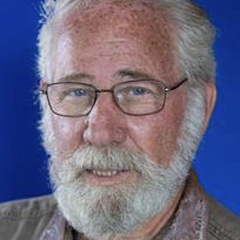 An older man with white hair, a full white beard, and mustache is wearing glasses. He has light skin and is dressed in a brown shirt. Against a solid blue background, Steve Boockvor looks directly at the camera with a gentle expression.