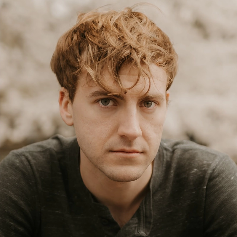 A young man with tousled light brown hair and fair skin looks directly at the camera with a neutral expression. He is wearing a dark gray shirt outdoors; the softly blurred, light-colored background was photographed by Phoebe Eskovitz.