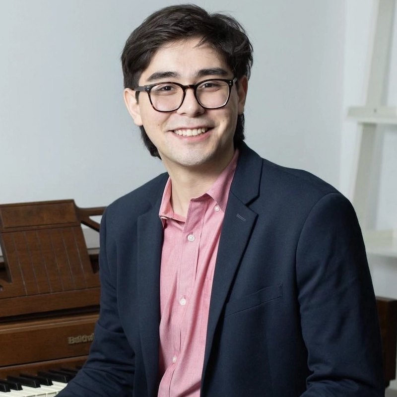 James Brandfonbrener, a young man with dark hair and glasses, smiles at the camera. He wears a dark blazer over a light pink shirt and is seated in front of an upright piano in a bright, modern room with white walls.