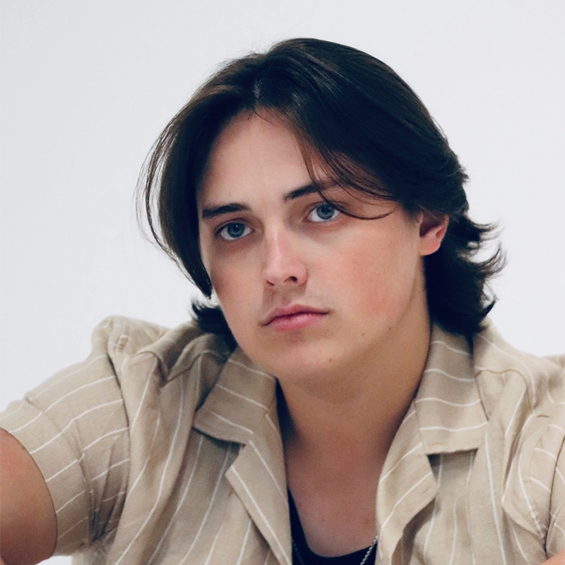 Phoebe Eskovitz, a young person with medium-length dark brown hair and blue eyes, looks directly at the camera with a neutral expression. They wear a beige shirt with white vertical stripes and sit against a plain white background.