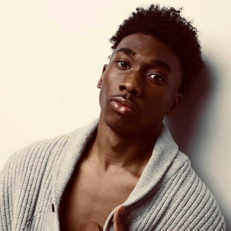 A young Black man with short curly hair, resembling Seth Remington, gazes at the camera with a neutral expression. He wears a light gray, ribbed cardigan draped open over his shoulders, exposing part of his chest against a softly lit background.
