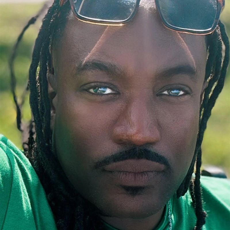 A close-up of a person with dark skin, blue-green eyes, a mustache, and goatee. They have long braided hair and are wearing sunglasses on their head and a green shirt. Sunlight is creating highlights on their face and background is blurred greenery.