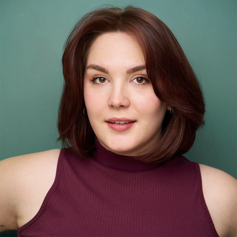 Leila Bruske, a woman with fair skin and shoulder-length brown hair, poses in front of a teal background. She wears a sleeveless, high-neck burgundy top, looking directly at the camera with a slight smile and natural makeup.