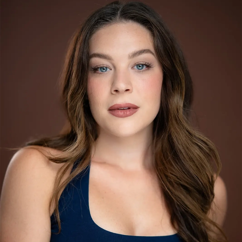 A woman with long, wavy brown hair and blue eyes gazes at the camera. She wears a navy blue top and natural makeup with bold brows and mauve lipstick. The softly blurred plain brown background highlights her face and upper shoulders, drawing attention to her striking resemblance to Courtney Burnett.
