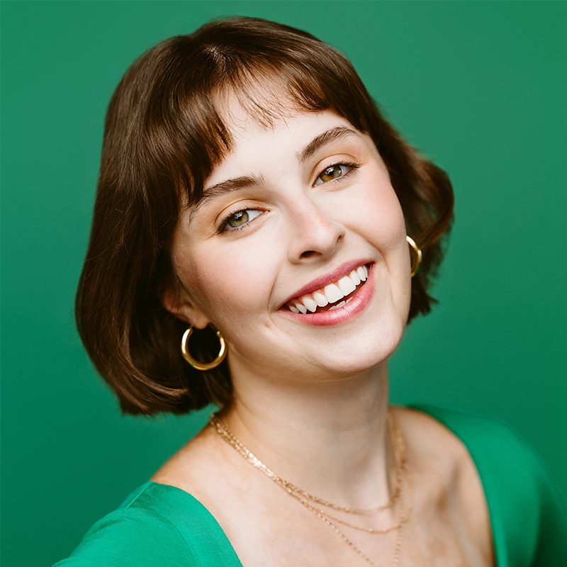 Phoebe Eskovitz, a young woman with short brown hair and bangs, smiles warmly. She wears gold hoop earrings, layered necklaces, and a green top that matches the solid green background, her head tilted slightly in a natural, friendly expression.