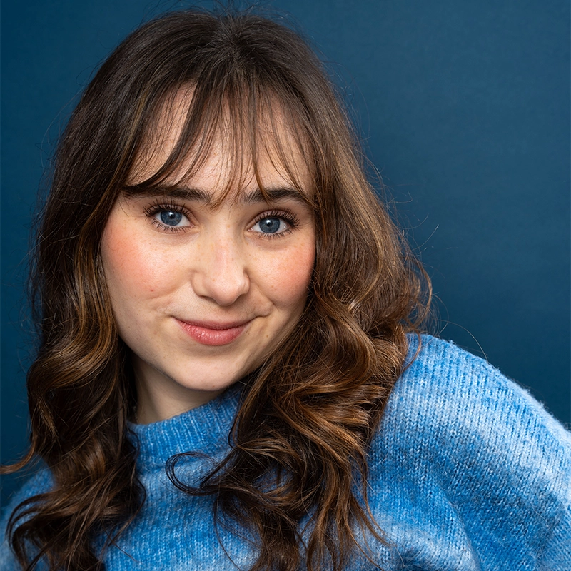 Olivia Luzquinos, a young woman with long wavy brown hair and blue eyes, smiles softly at the camera. She wears a light blue sweater and stands in front of a plain dark blue background, her cheeks showing a natural rosy blush.