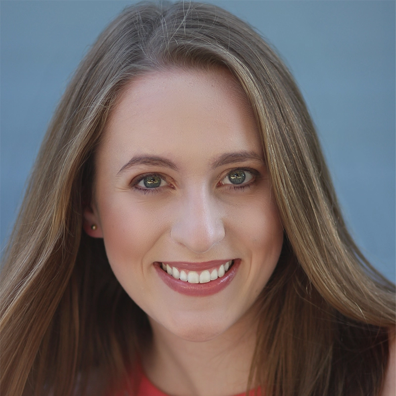 Caroline Howard, a young woman with long, straight light brown hair, smiles warmly at the camera. She has green eyes, light skin, and wears subtle makeup. The background is a soft blue, and she is dressed in a coral-colored top.