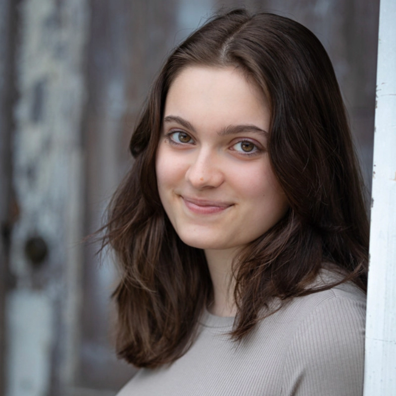 Phoebe Eskovitz, a young woman with shoulder-length brown hair and light skin, stands near a weathered wooden door, smiling softly at the camera. She wears a light beige ribbed top against a backdrop of faded paint and rustic textures.