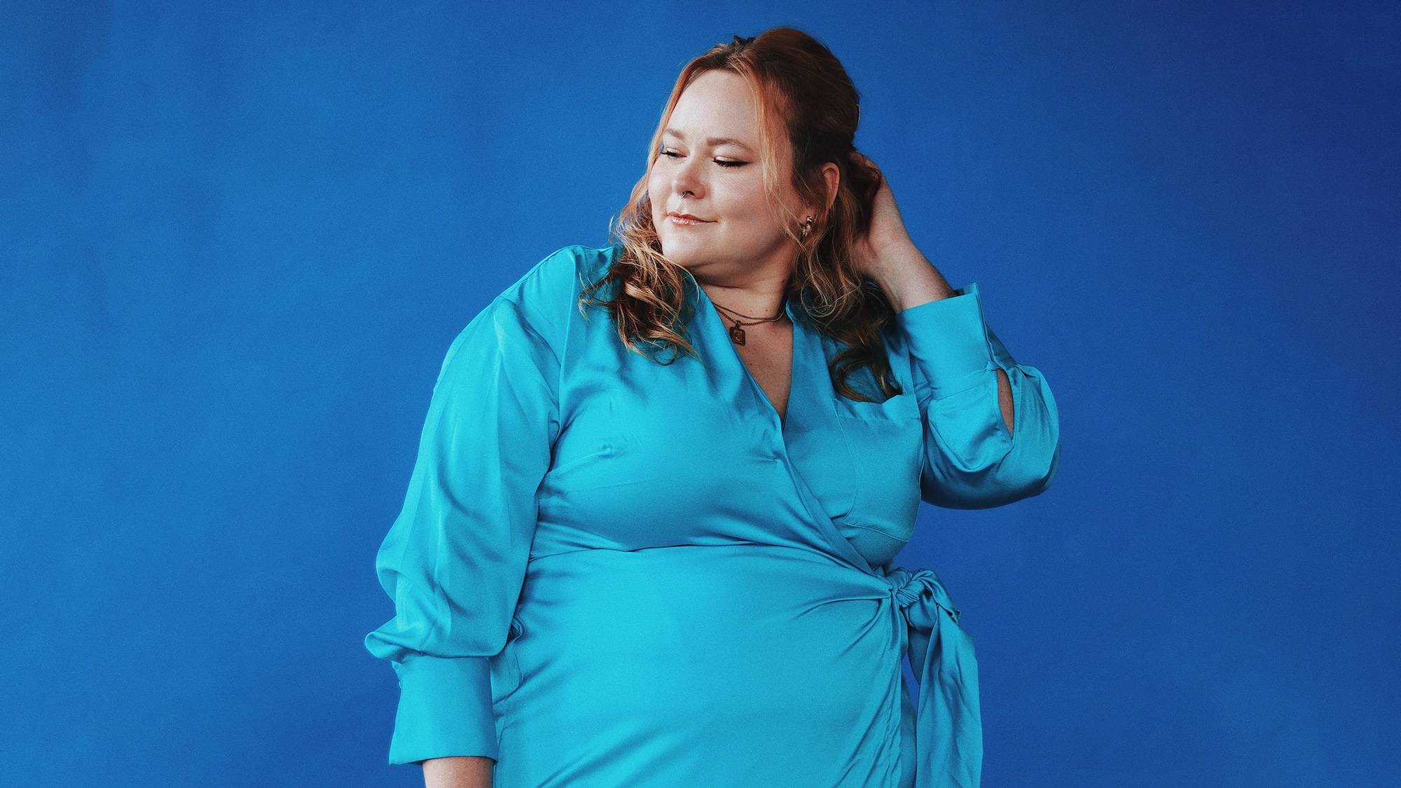 Cassi Mikat site image 25 A woman stands against a solid blue background, wearing a vibrant blue wrap dress from the Sing Loud collection by Cassi Mikat. She has light skin, long wavy hair, and looks down with a slight smile, one hand raised to touch her hair near her neck.