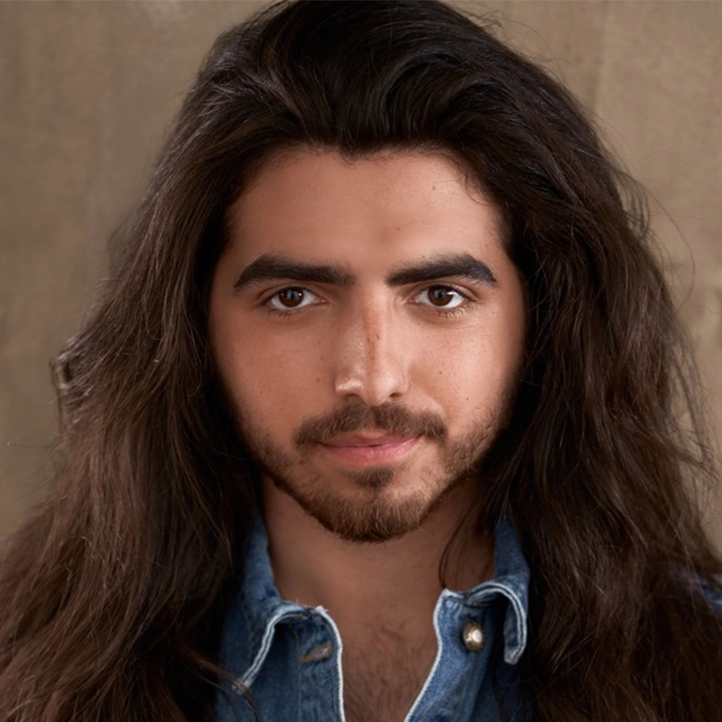 A young man with long, wavy dark hair and a trimmed beard looks directly at the camera. He wears a blue denim shirt with the top buttons open, standing against a warm-toned wall—a style reminiscent of Marielle Utayde’s portrait work.