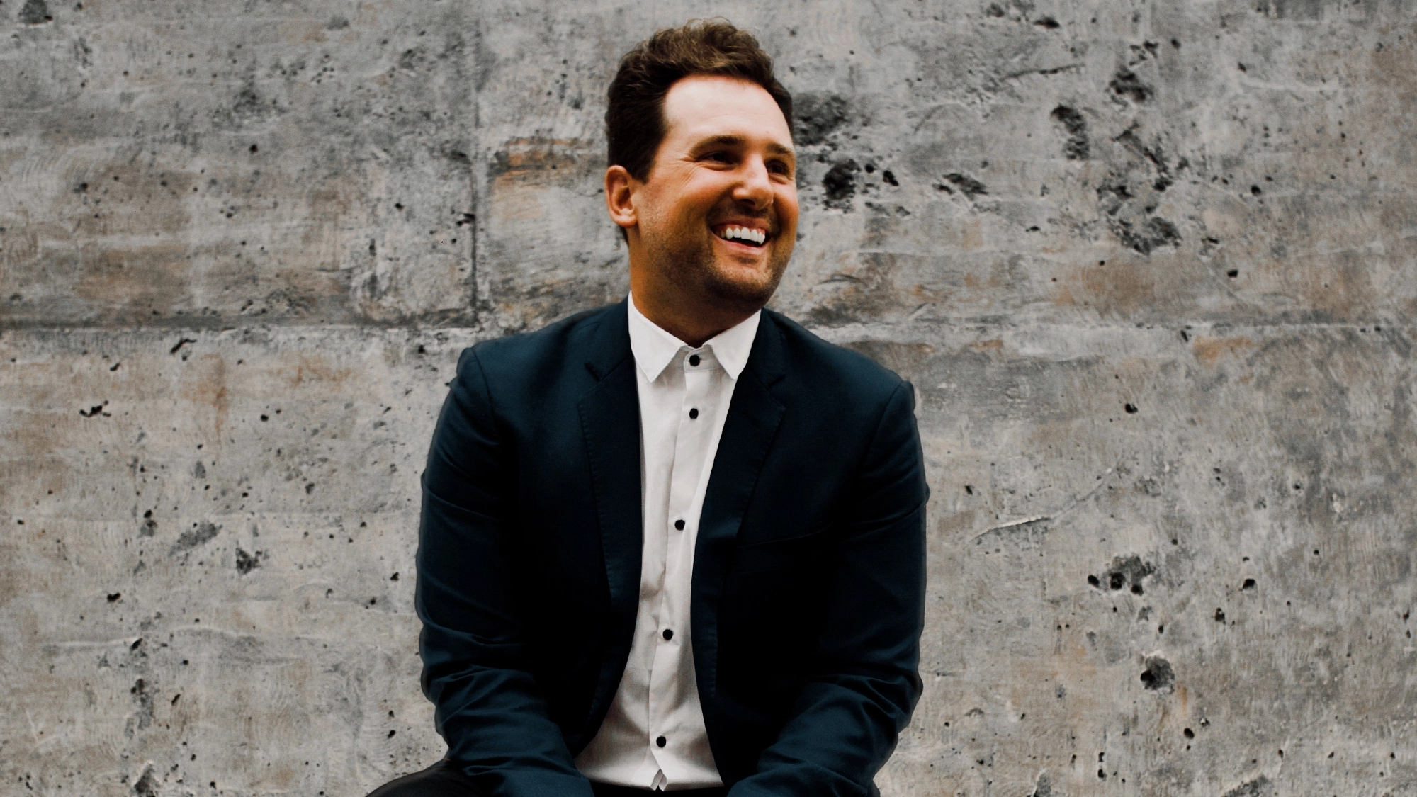 Chris Pinnella site image 26 A man in a dark suit and white shirt sits smiling and looking to his left. He has short brown hair and a trimmed beard. The modern, casual scene recalls Chris Pinnella at ease before performing with a chamber orchestra.