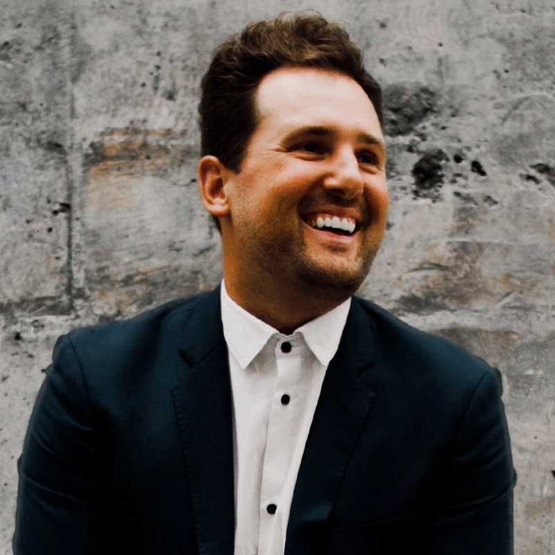 A man with short brown hair and a trimmed beard smiles while looking to the side. He wears a dark suit jacket over a white shirt with black buttons. The background is a textured gray stone wall. The lighting is soft and natural.