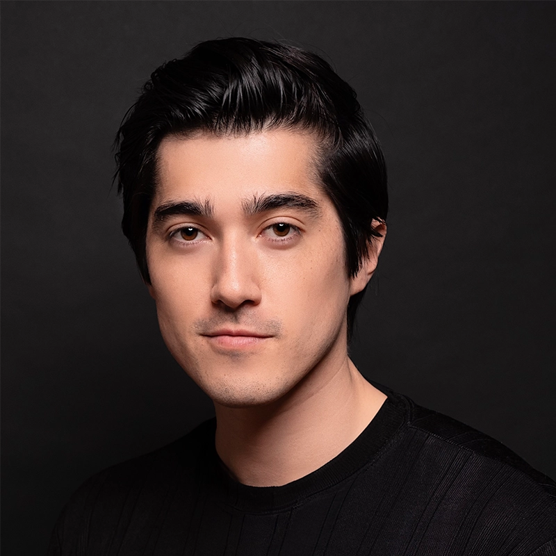 A young man with fair skin and short, dark hair poses against a plain black background. He has brown eyes, a clean-shaven face, and wears a black crew-neck shirt. Chris Ruetten’s neutral, confident expression faces the camera directly.