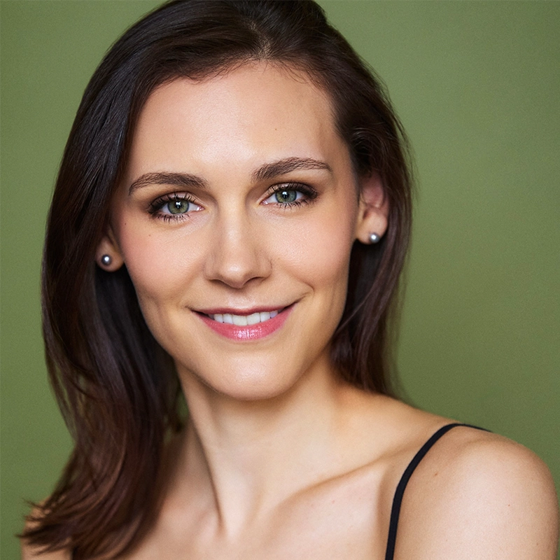 Adriana Ellis, a woman with light skin, straight brown hair, and blue eyes, smiles softly at the camera. She wears pearl earrings and a thin-strap black top. The bright, even lighting highlights her clear complexion against an olive green background.