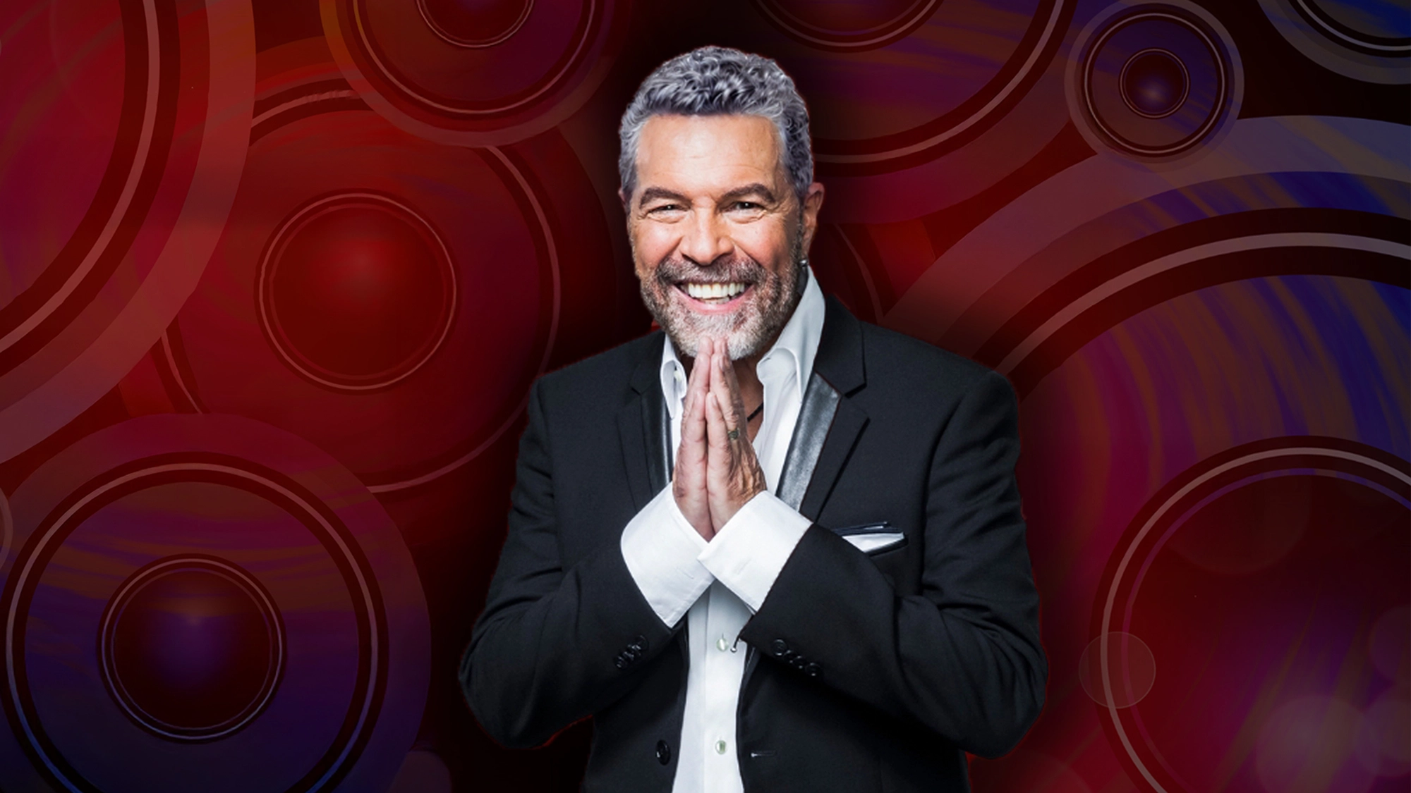 Clint Holmes - Show Art A smiling man with gray hair and beard, wearing a black suit and white shirt, stands with hands pressed together in front of him. The vibrant background of red circles and rings hints at a performance by Clint Holmes or Joni.