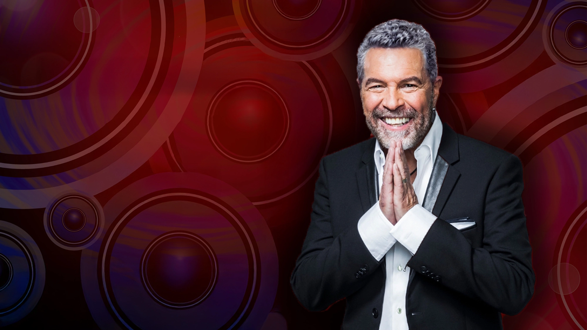 A smiling man with gray hair and beard, dressed in a black suit with a white shirt, stands with palms pressed together in front of his face. The background features vibrant red and purple concentric circles with a glowing effect, evoking Clint Holmes’ stage presence.