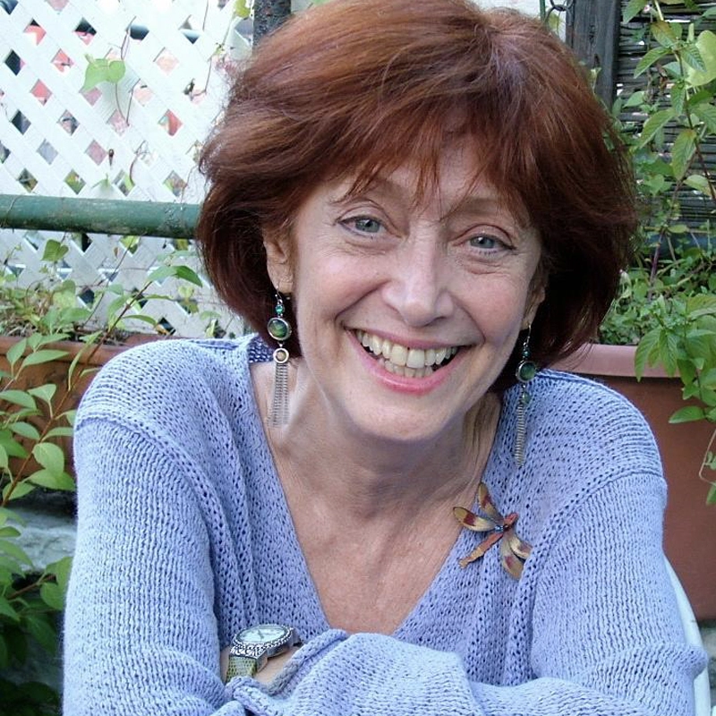 Margery Cohen, a smiling older woman with short reddish hair, wears a light purple sweater, green earrings, and a dragonfly brooch as she sits in a garden setting with lush plants and a white lattice fence behind her.