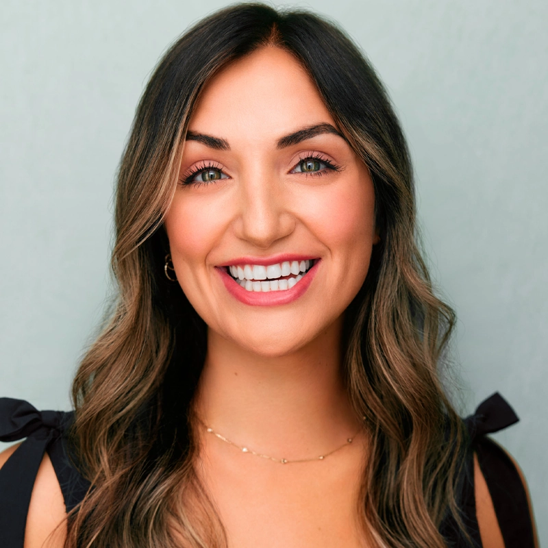 A woman with long, wavy brown hair and highlights smiles at the camera. She wears a black top with tied shoulder straps and a delicate gold necklace. The background is a soft, light teal color. Her makeup is natural with a bold pink lipstick.