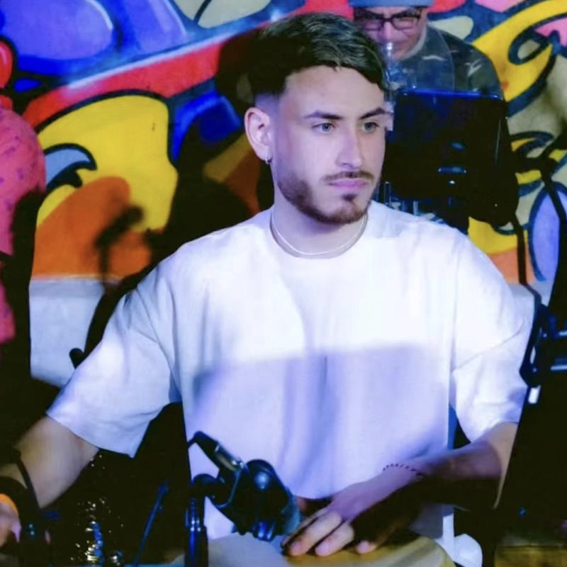 A young man with short, light brown hair and a trimmed beard, resembling Facundo Colman, sits at a table with microphones and equipment. He wears a white t-shirt and looks slightly to the side. Behind him is a colorful graffiti wall and a person with a camera.