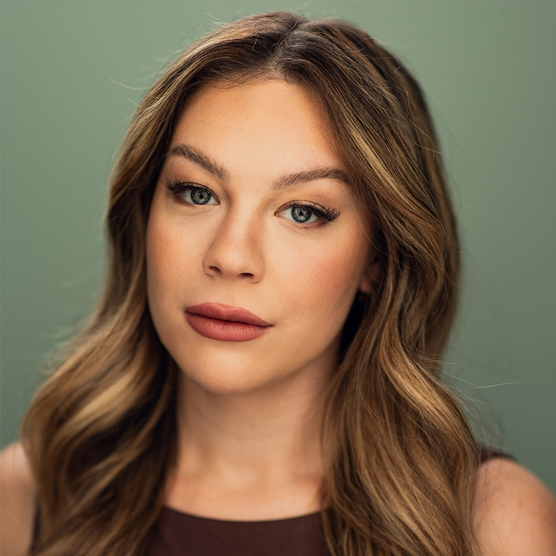 Courtney Burnett, with long, wavy light brown hair and green eyes, looks at the camera, wearing natural makeup with defined eyebrows and muted pink lipstick. She is dressed in a brown sleeveless top against a plain sage green background.