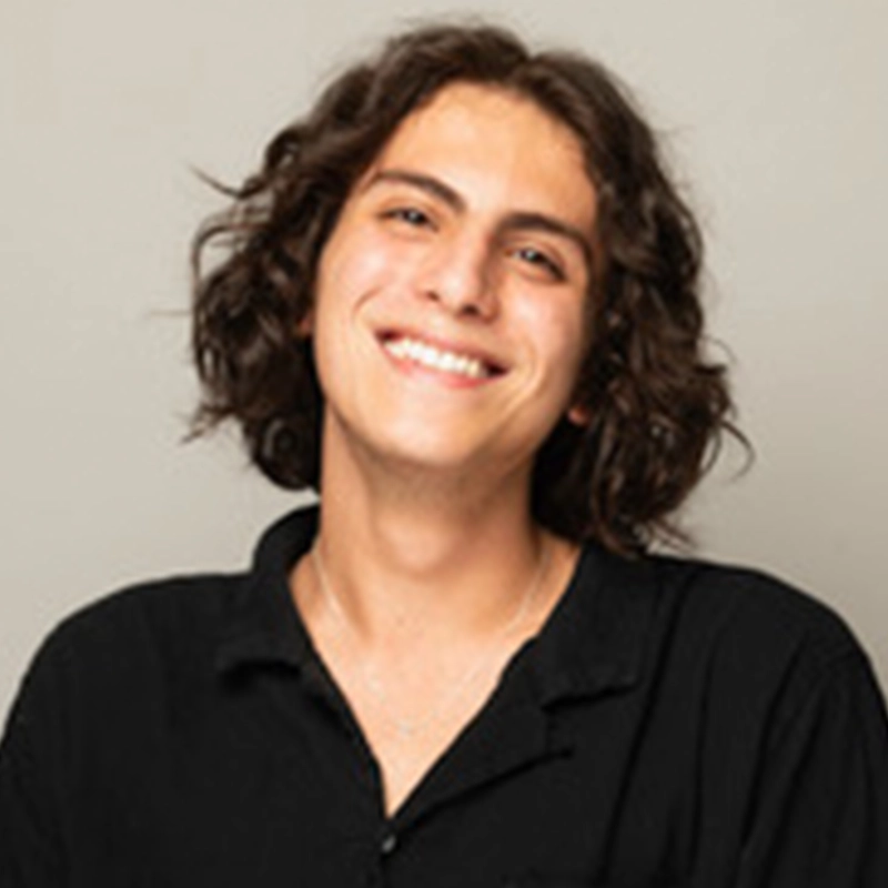A young person with shoulder-length curly brown hair smiles warmly at the camera. They are wearing a black button-up shirt and a thin necklace. The plain, light background highlights their friendly, approachable vibe—perfect for Marguerite A. Boone’s profile.