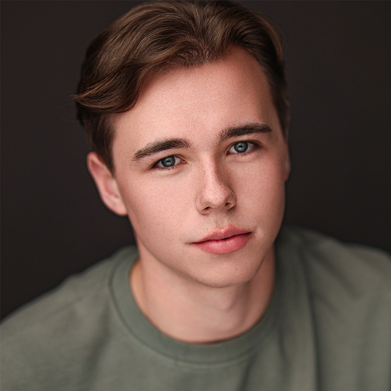 A young man with fair skin, short brown hair, and blue eyes gazes softly at the camera. Wearing a sage green crew-neck shirt, he poses against a dark backdrop; the soft lighting highlights his clear complexion in classic Phoebe Eskovitz style.