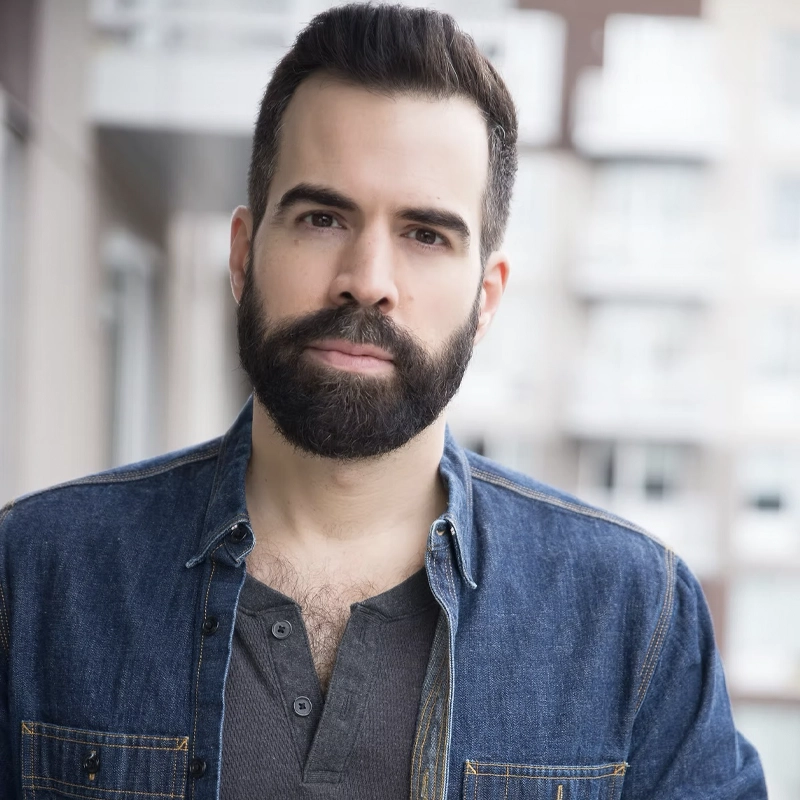 Christopher DeProphetis, a man with dark hair and a full beard, stands on a balcony in a denim shirt over a buttoned grey undershirt. He looks directly at the camera with a slight smile; behind him are blurred modern apartment buildings.