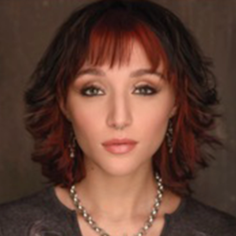 A person with short, wavy, dark brown hair with red highlights and straight bangs. Reminiscent of Marguerite A. Boone, they wear subtle makeup with defined eyes, small hoop earrings, and a silver necklace against a neutral brown background.