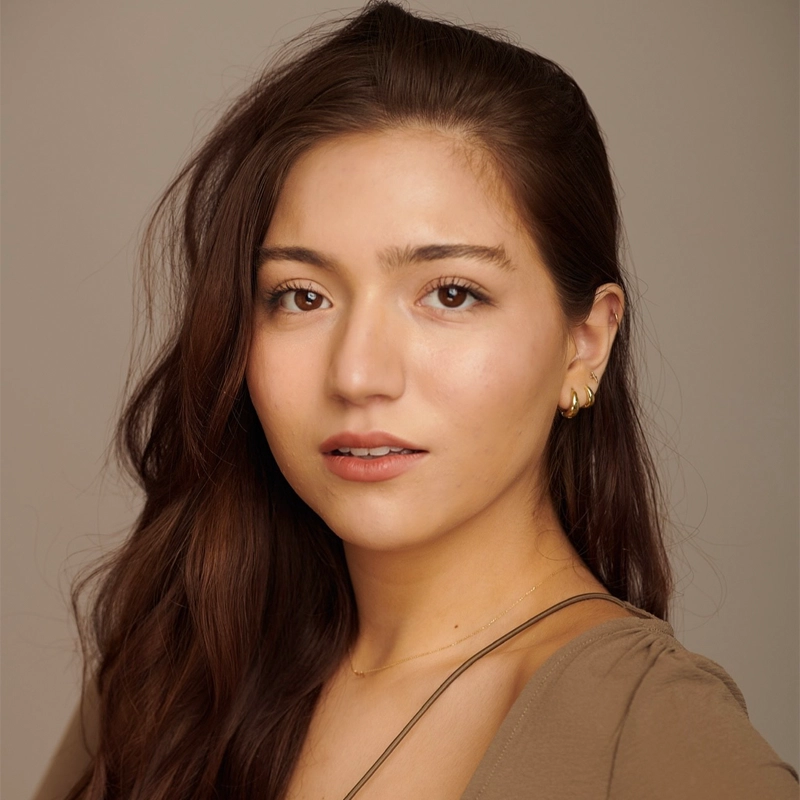 A young woman with long, wavy brown hair and light skin looks at the camera. She wears a beige top, gold hoop earrings, and a delicate gold necklace by Mar Di Meglio. The plain background creates a soft, professional portrait atmosphere.