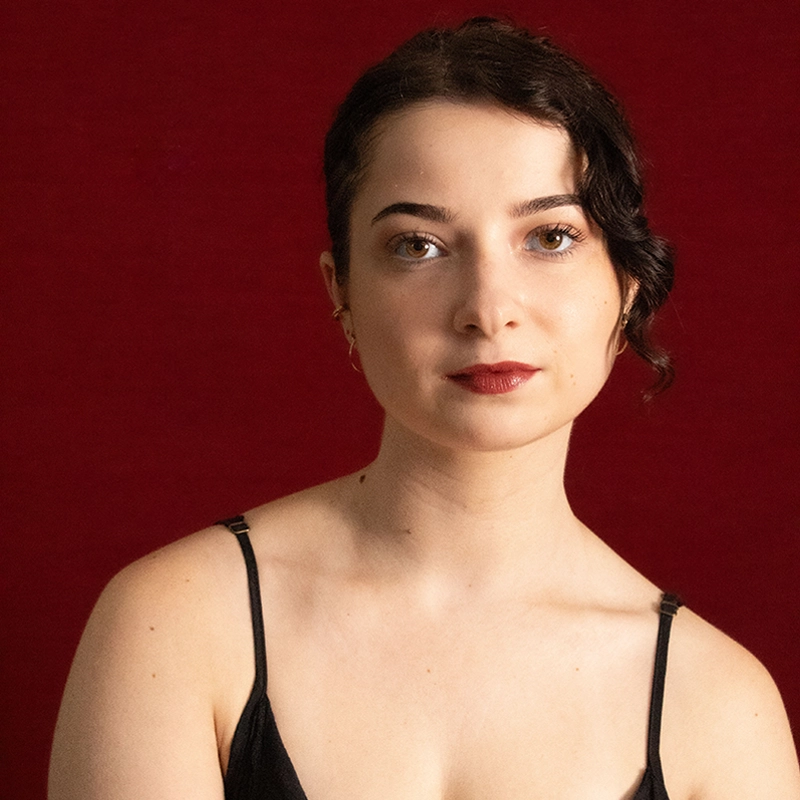 A young woman with fair skin and short, dark curly hair poses against a deep red background. Charlie Valdez wears a black spaghetti strap top and subtle makeup with bold red lipstick, gazing calmly at the camera with a neutral expression.