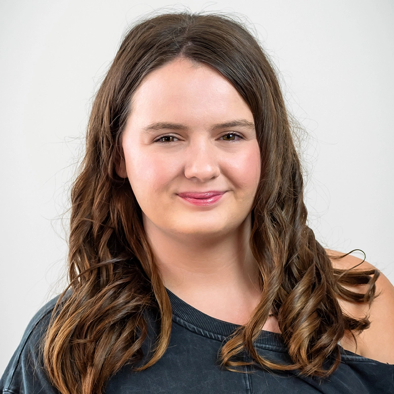A young woman with long, wavy brown hair smiles softly at the camera. She is wearing a dark, off-the-shoulder top and has light skin. The plain, light background keeps the focus on her face—an inviting portrait by Dylan McEachern.