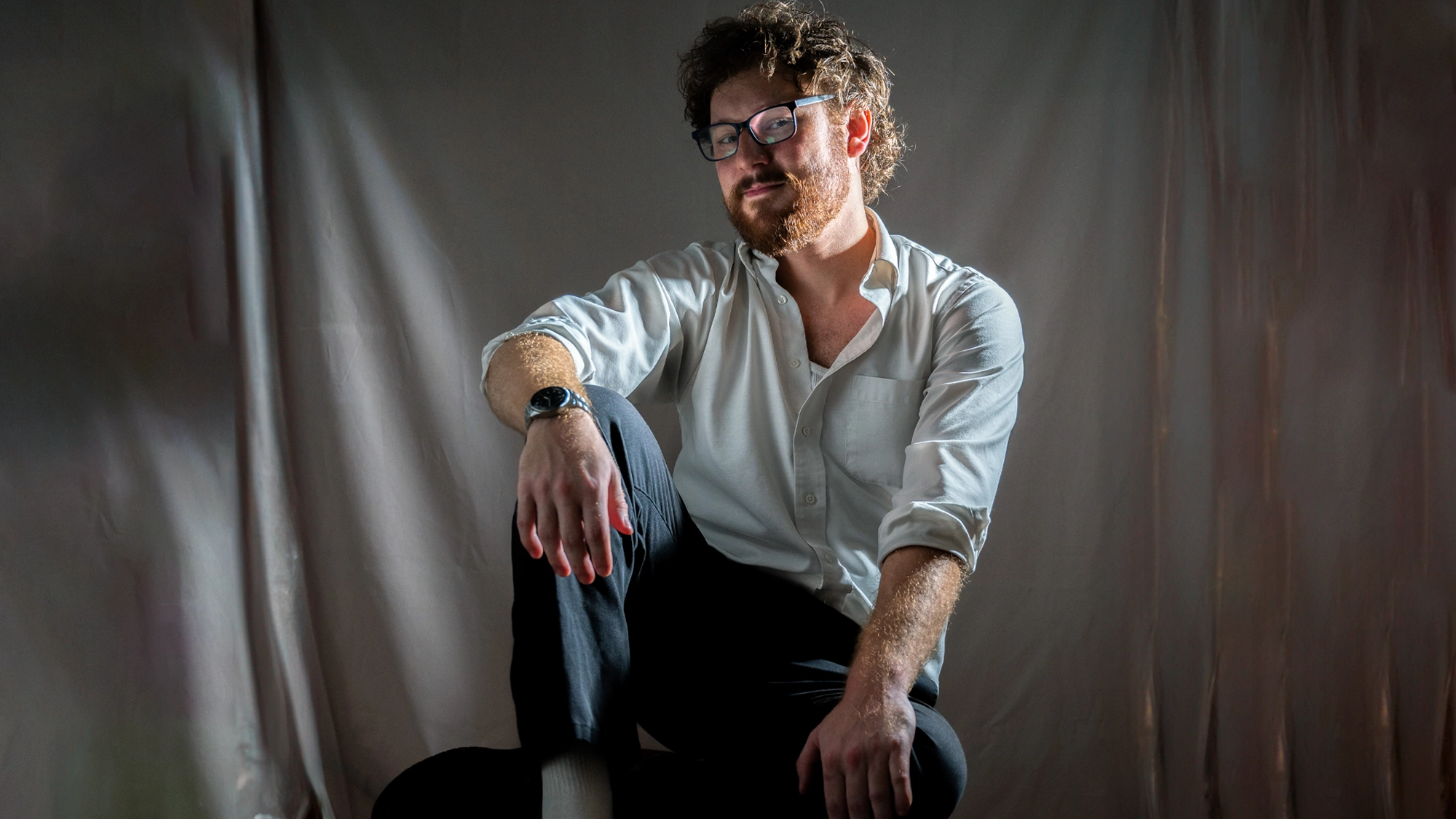 A man with curly hair and glasses—reminiscent of Eddie Bean—sits on a chair, resting one arm on his knee. He wears a white button-down shirt and dark pants. Soft, dramatic lighting highlights his face and relaxed pose against a neutral fabric backdrop.