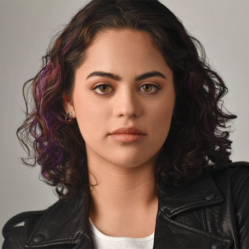 A young woman with wavy dark hair and subtle purple highlights looks directly at the camera. Reminiscent of Camelia Elías, she has light skin, brown eyes, and wears a white top under a black leather jacket against a softly illuminated neutral background.