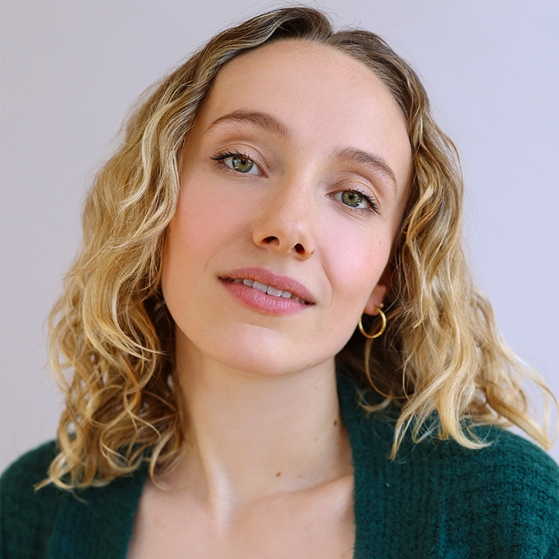 Amanda Harris, a woman with light curly blonde hair, fair skin, and green eyes, looks at the camera with a gentle expression. She wears a green textured top and small gold hoop earrings against a plain, softly lit background.