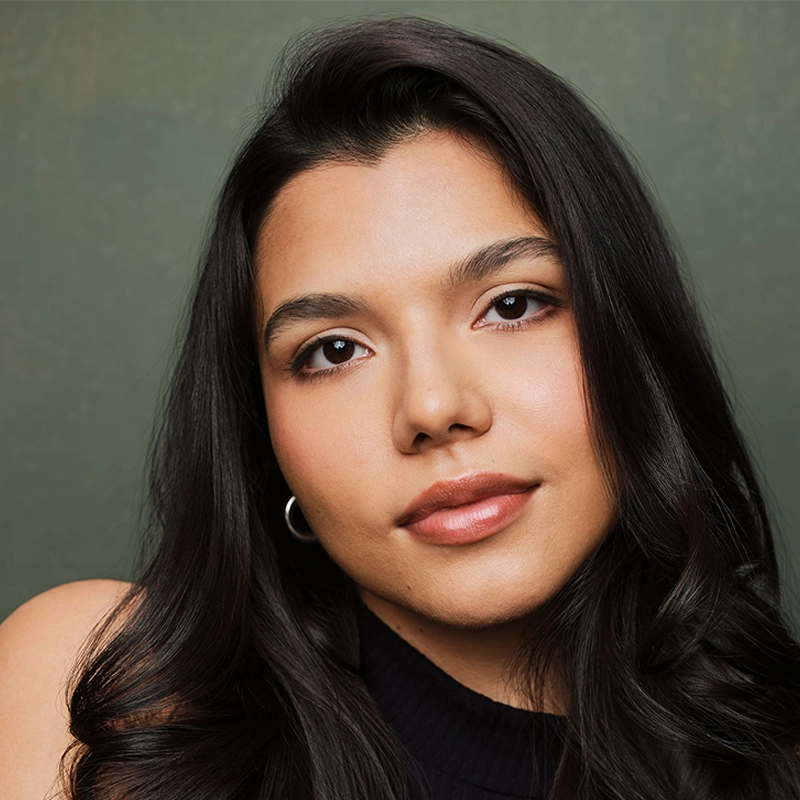 Elizabeth Raquel Ramirez, a young woman with long dark hair and smooth skin, looks at the camera with a gentle expression. She wears a sleeveless black top and small hoop earrings, posing in front of a muted green background.