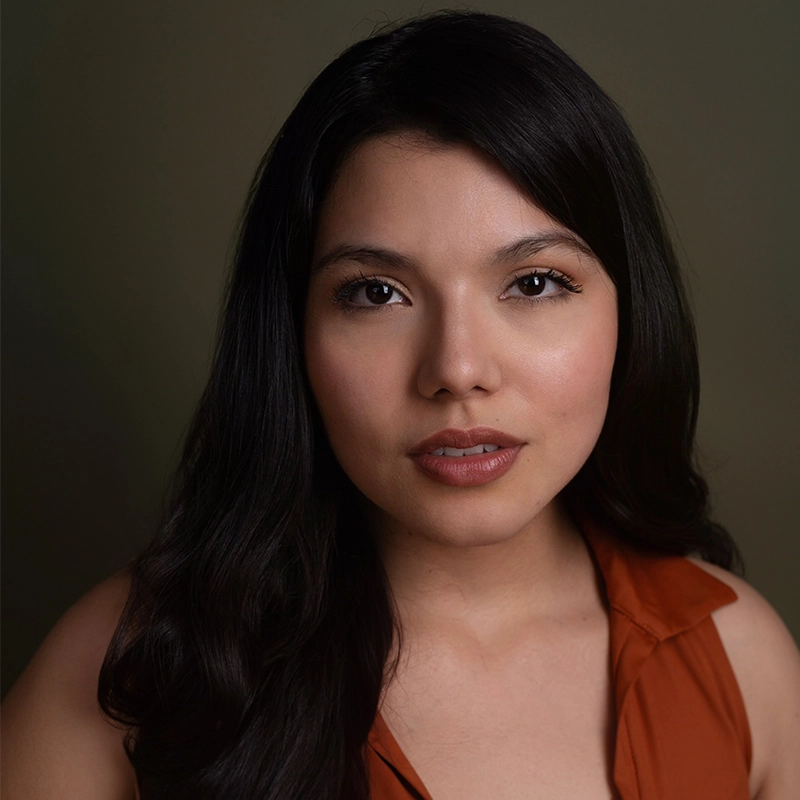 Elizabeth Raquel Ramirez, with long, dark hair and light brown skin, stares directly at the camera. She wears a sleeveless, rust-colored top and natural makeup with defined brows and soft pink lips against a plain dark background.