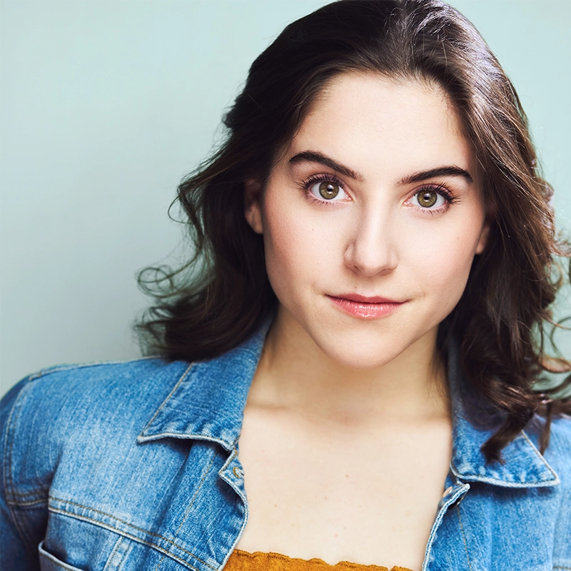 Adriana Ellis, a young woman with wavy dark brown hair and fair skin, looks directly at the camera, wearing a light denim jacket over a mustard yellow top. She has full eyebrows, hazel eyes, and natural makeup against a soft pale green background.