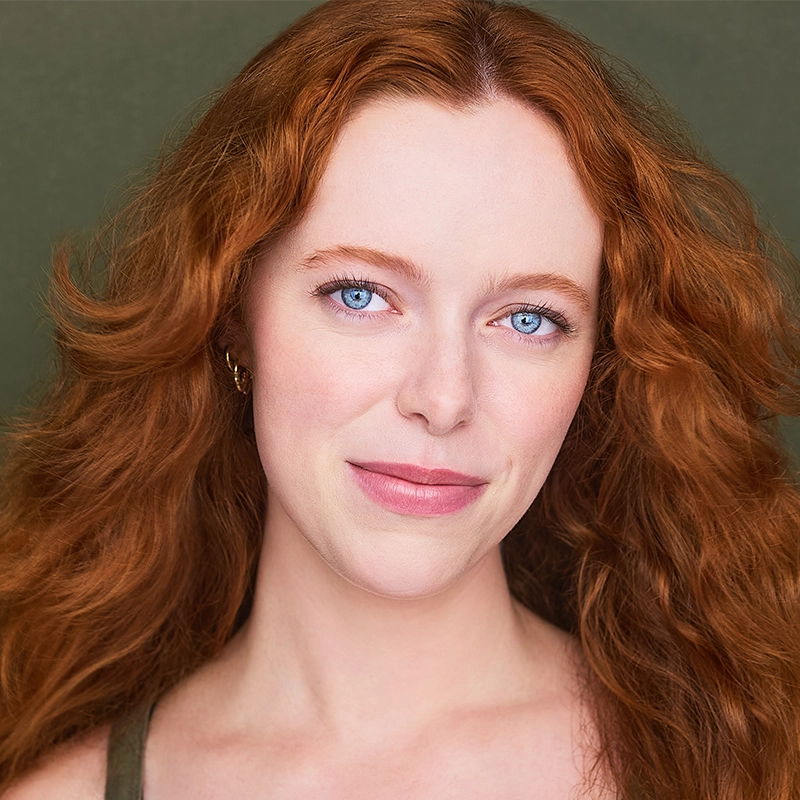 Elizabeth Raquel Ramirez, a woman with long, wavy red hair and striking blue eyes, looks directly at the camera with a slight smile. She has fair skin, natural makeup, and is set against a muted green background.