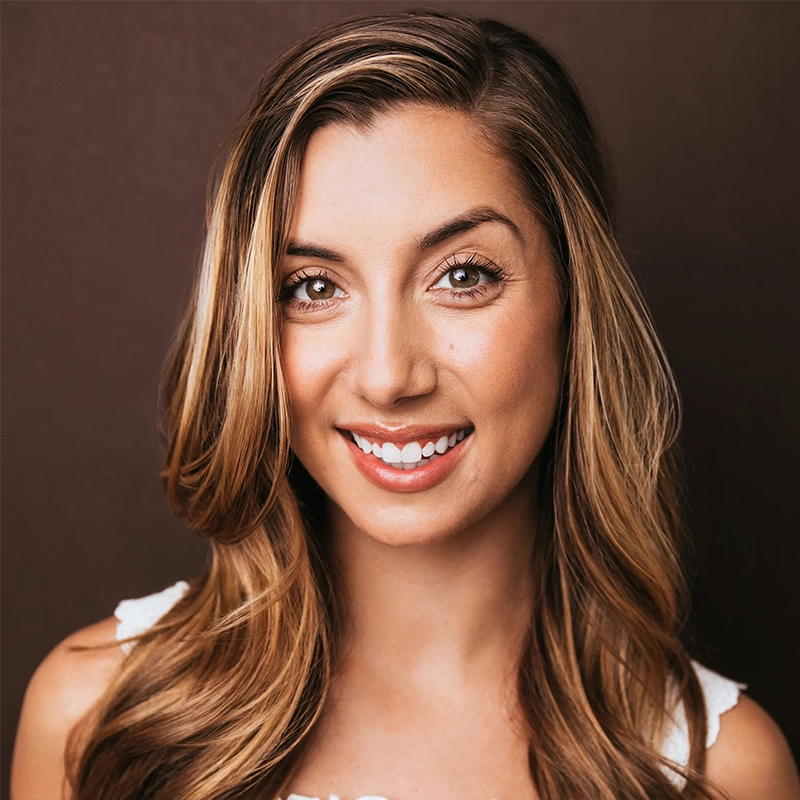 A young woman with long, wavy, light brown hair smiles at the camera. She has fair skin, brown eyes, and is wearing a white sleeveless top. The dark brown background contrasts with her friendly, confident expression in this Josiah Jennings portrait.