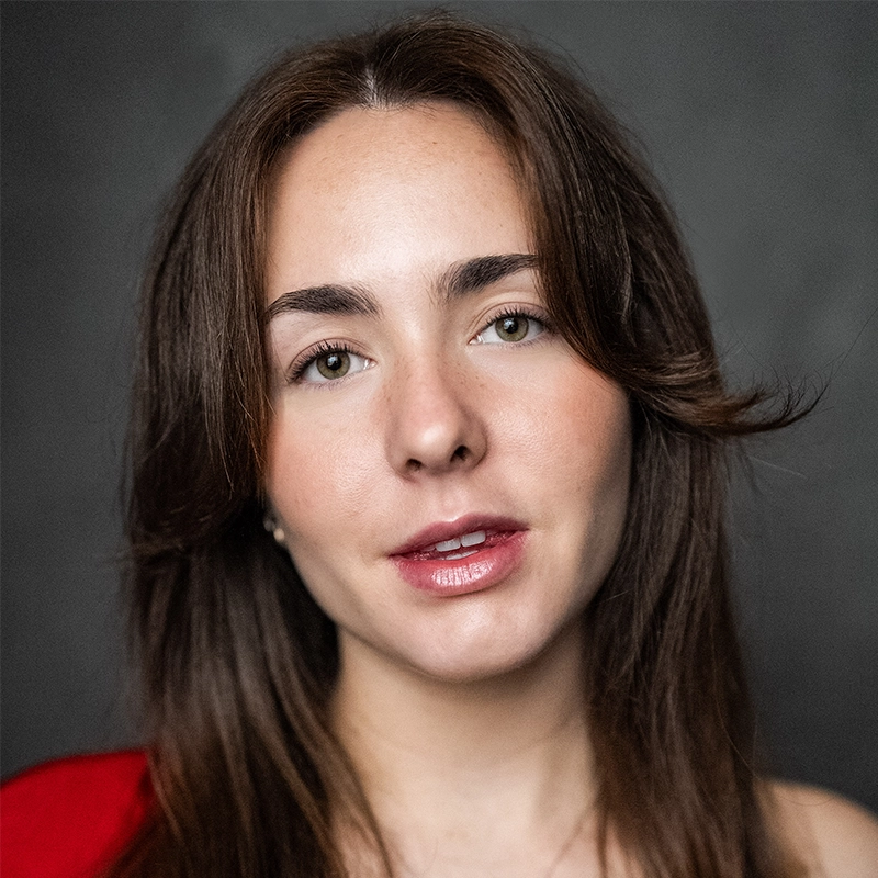 A young woman with fair skin, long brown hair, and hazel eyes faces the camera. Emma Wallace wears natural makeup with glossy lips and slightly arched brows. She is dressed in a red top against a soft, dark gray background.